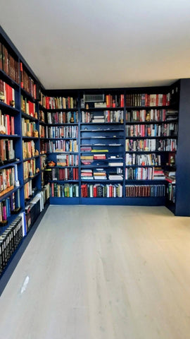 A large home library with dark navy blue bookshelves lining the walls, filled with books, and a light-colored wood floor.