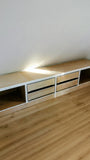 A low wooden storage bench or TV stand with open shelving and two drawers, shown in a room with light wood flooring and a plain wall.