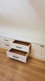 White built-in drawers with black handles, with two lower drawers pulled open to show the wood interior, set against a white wall on a light wood floor.