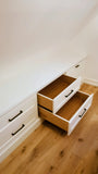 White built-in drawers with black handles on a sloped ceiling, with two middle drawers pulled open to show a light wood interior, set on a light wood floor.
