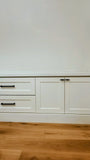 White built-in cabinetry with two drawers on the left and two cabinet doors on the right, featuring slim black metal handles, set against a white wall and light wood flooring.