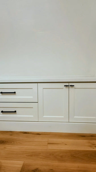 White built-in cabinetry with two drawers on the left and two cabinet doors on the right, featuring slim black metal handles, set against a white wall and light wood flooring.