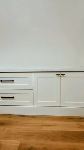 White built-in cabinetry with two drawers on the left and two cabinet doors on the right, featuring slim black metal handles, set against a white wall and light wood flooring.