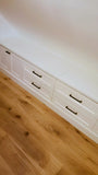 White built-in storage drawers with black metal handles on a wooden floor against a white wall.